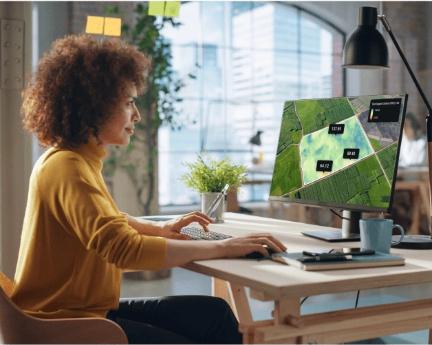 A woman sits in front of a computer display and analyzes SOC levels on her fields remotely with the EOSDA Carbon solution